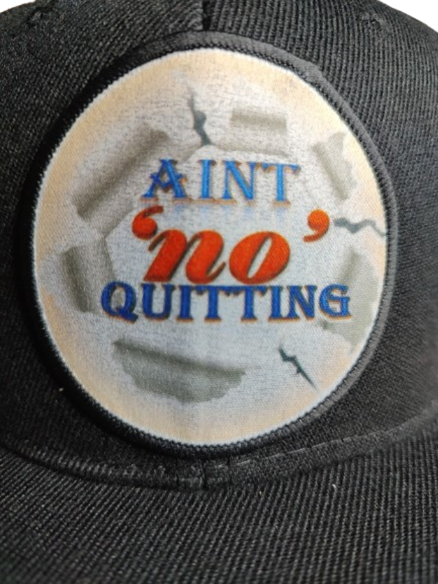 A black flat-brim baseball cap featuring a circular white patch on the front. The patch has a ripped paper effect around the edges and displays the motivational phrase "AIN'T 'NO' QUITTING" in blue and orange text. This cap is part of a never quit gift bundle, ideal for a fitness motivation gift set or empowerment apparel.
www.justsaynpromos.com